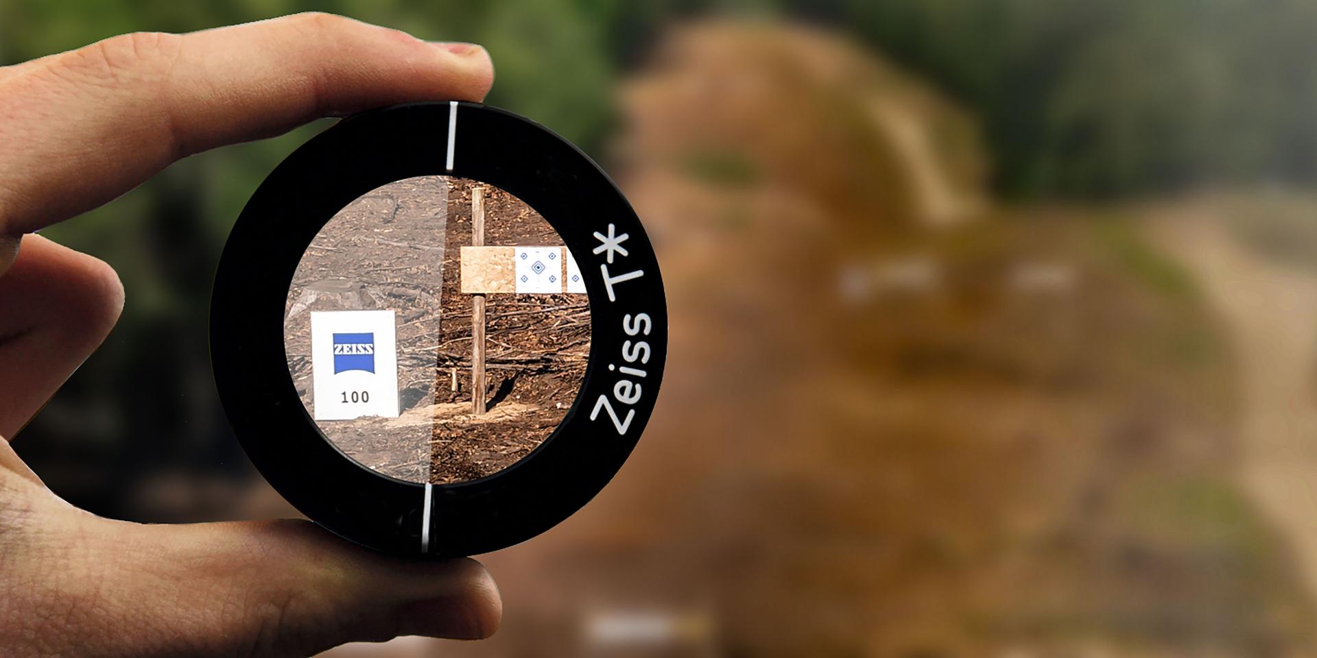 A hand holds a ZEISS lens filter showing a clear, detailed view of a forest path through the lens, contrasting with the blurred background.
