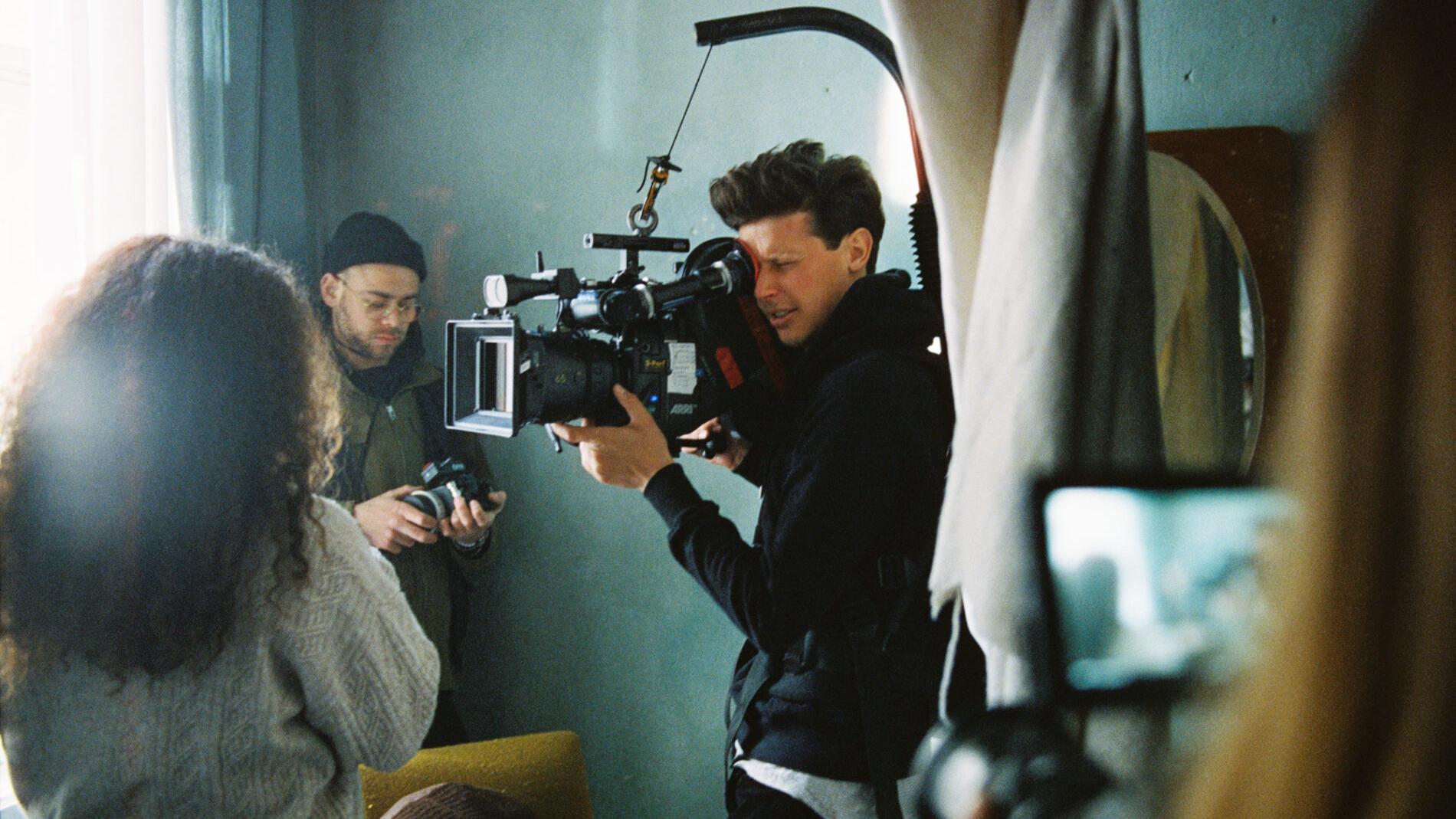 A cameraman operates a professional film camera equipped with a ZEISS lens during a video shoot with a small production crew.