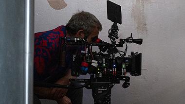 A camera operator is crouched behind a professional cinema camera rig equipped with ZEISS lenses, positioned on wooden boxes in front of a textured wall.