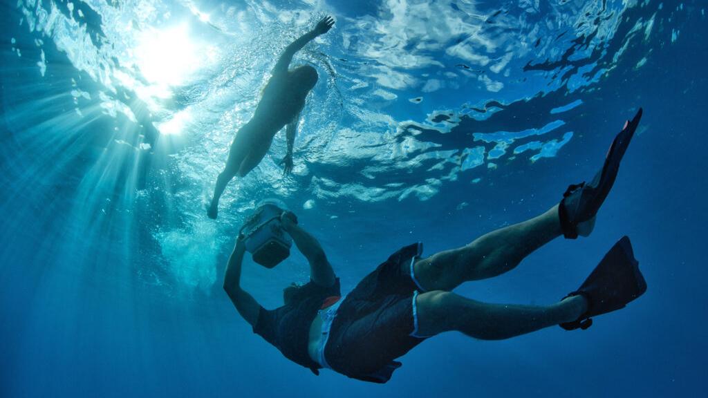 Two snorkelers swim underwater in clear blue ocean light with sunlight filtering through the surface, captured in sharp detail.