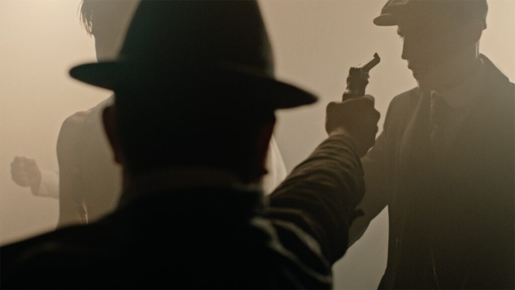 A dramatic, cinematic scene featuring three men in vintage clothing, with one man pointing a gun at another, captured in low light and atmospheric haze to showcase ZEISS lens performance.