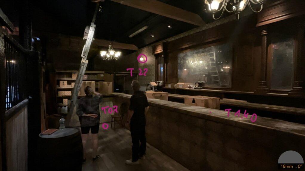 Two people stand in a dimly lit, rustic-style bar or tavern set, with lighting setup notes marked on the image and a ladder extended toward the ceiling, showcasing a ZEISS 18mm lens view.