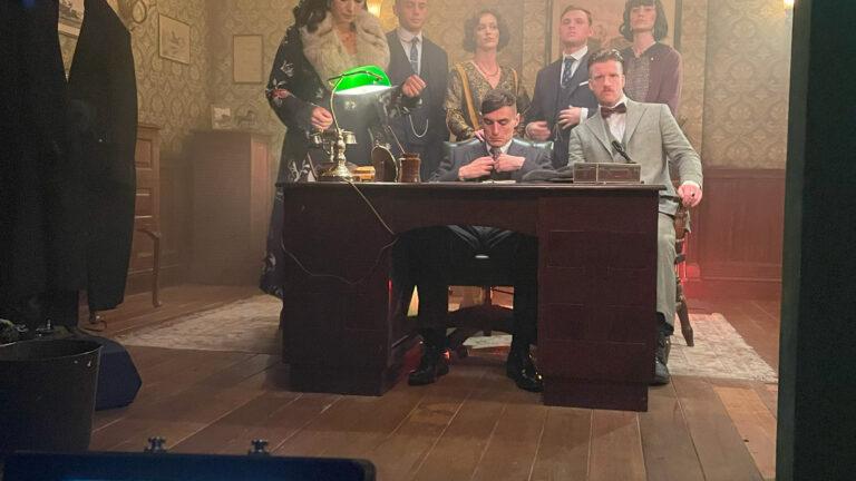 Behind-the-scenes view of a film set showing a group of actors in vintage attire posed around a desk, with a monitor in the foreground displaying the scene, captured using ZEISS lenses.