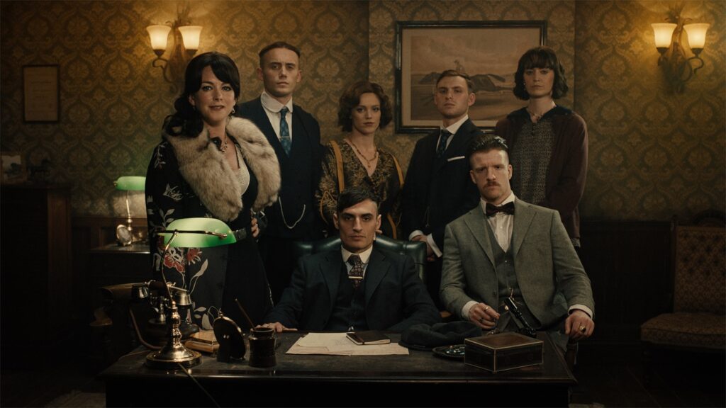 Seven people in vintage-style clothing pose formally in an old-fashioned office setting with retro wallpaper, wooden furniture, and a green-shaded desk lamp, showcasing the atmosphere in sharp detail and rich color as captured by ZEISS optics.