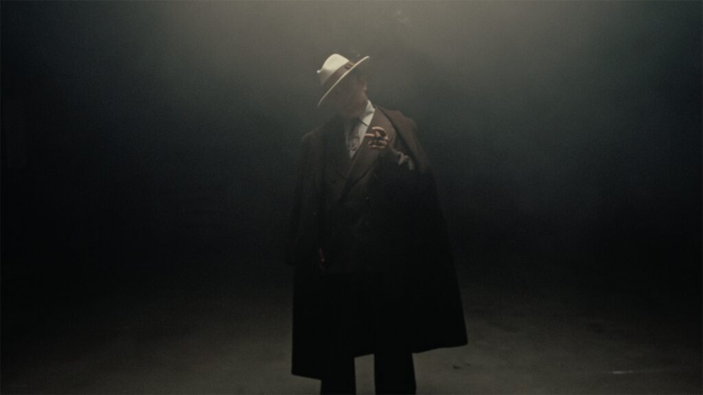 A man in a dark, foggy setting wears a white hat and formal coat, partially obscured by shadows and diffused light.