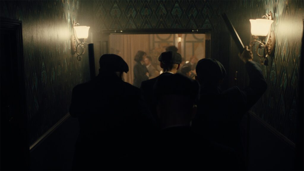 Four men in dark clothing and hats walk down a dimly lit hallway toward a lively, crowded room.