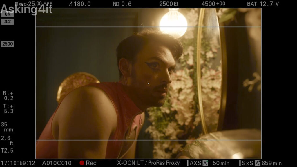 A man with theatrical makeup is framed in a dimly lit scene, looking into a mirror surrounded by flowers, as viewed through a professional camera monitor using ZEISS lenses.