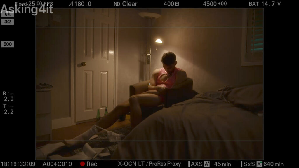A person sits alone in a dimly lit room on a chair next to a bed, captured through a professional film camera interface using ZEISS lenses.