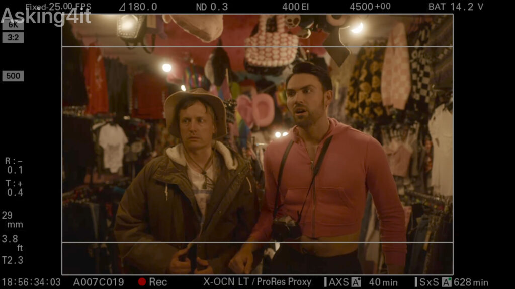 Two men stand inside a colorful, eclectic clothing store, captured through a camera monitor interface with ZEISS lens settings displayed.