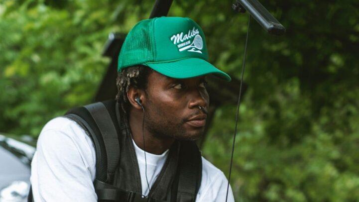 A camera operator kneels on grass while adjusting a professional cinema camera rig equipped with ZEISS lens components.