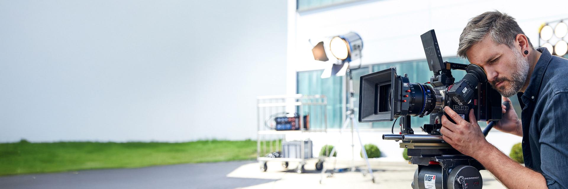 A man operates a professional video camera equipped with a ZEISS lens on an outdoor film set.