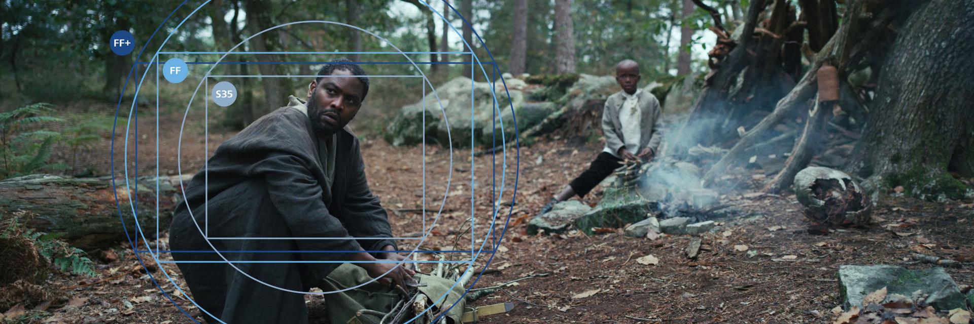 Two people are in a forest setting near a smokey campfire, with ZEISS lens interface graphics overlaid on the image.