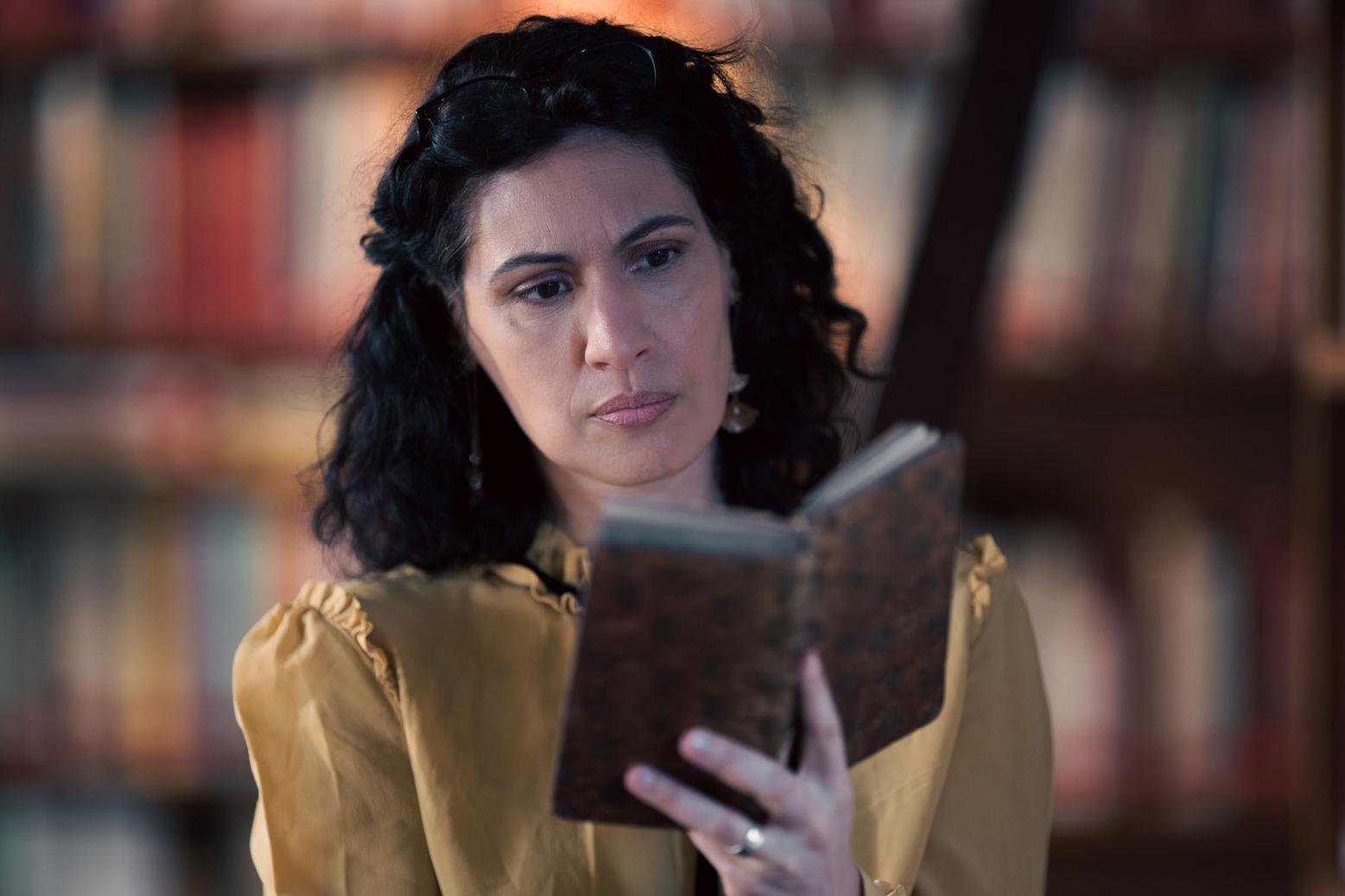 A woman stands in a historic library filled with bookshelves and long wooden tables covered in documents, viewed through a ZEISS lens.