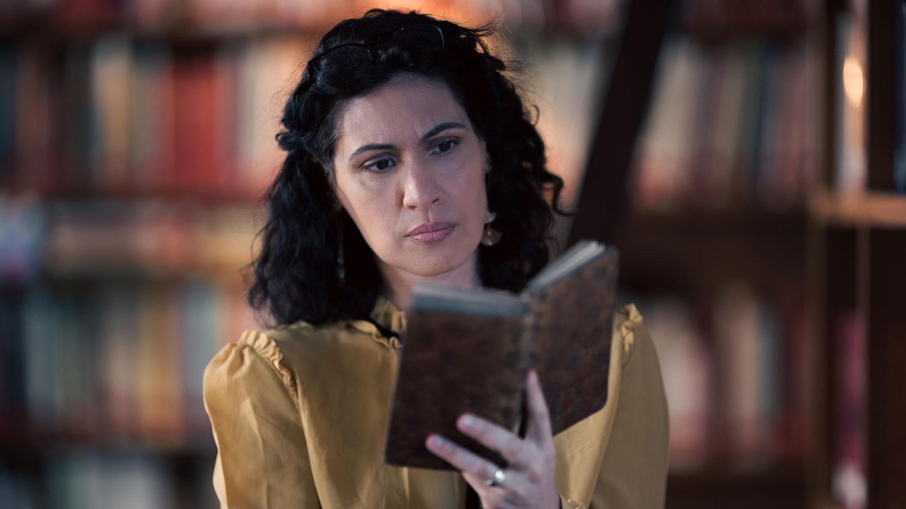 A woman stands in a historic library filled with bookshelves and long wooden tables covered in documents, viewed through a ZEISS lens.