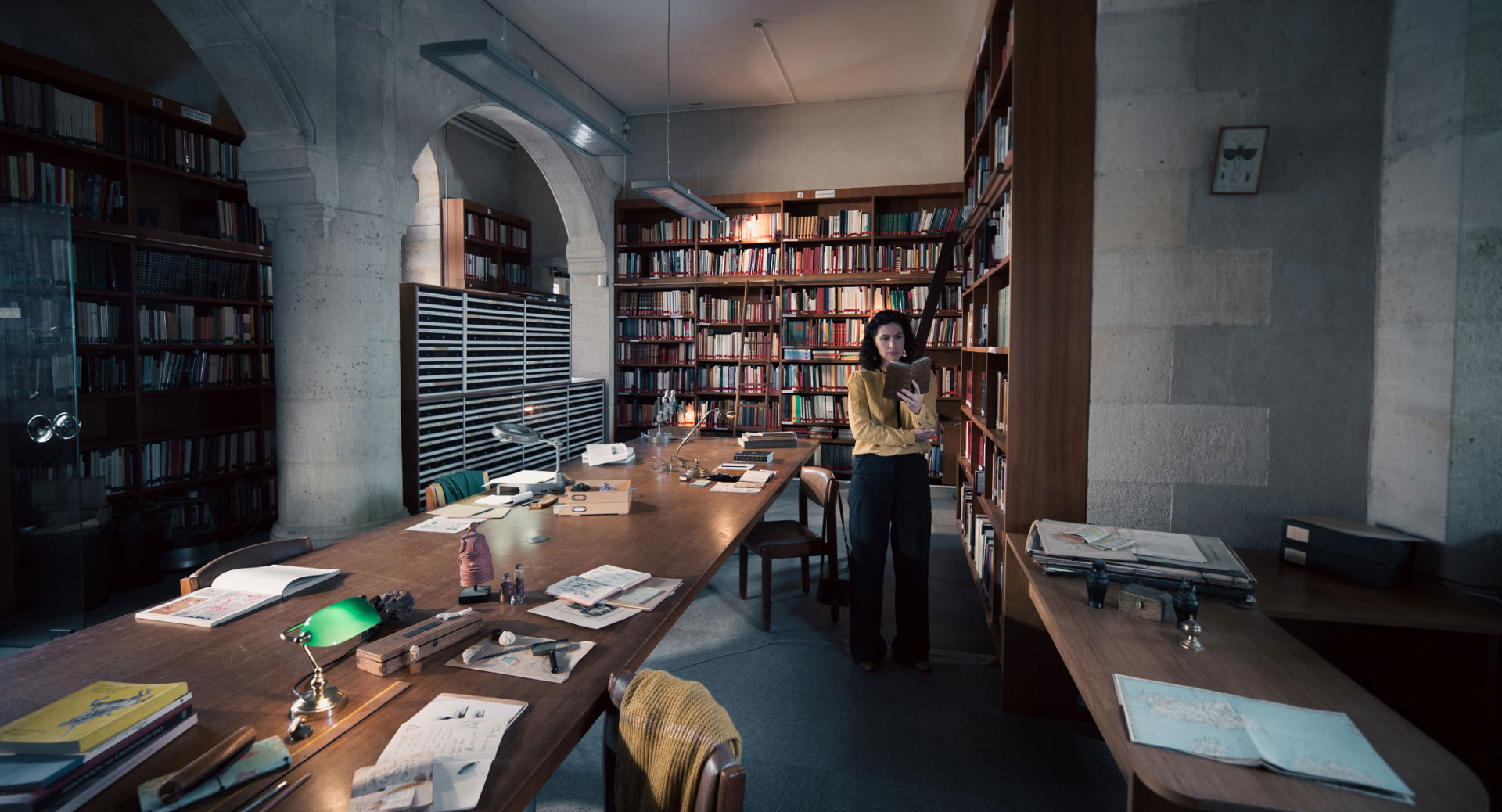 A woman stands in a historic library filled with bookshelves and long wooden tables covered in documents, viewed through a ZEISS lens.