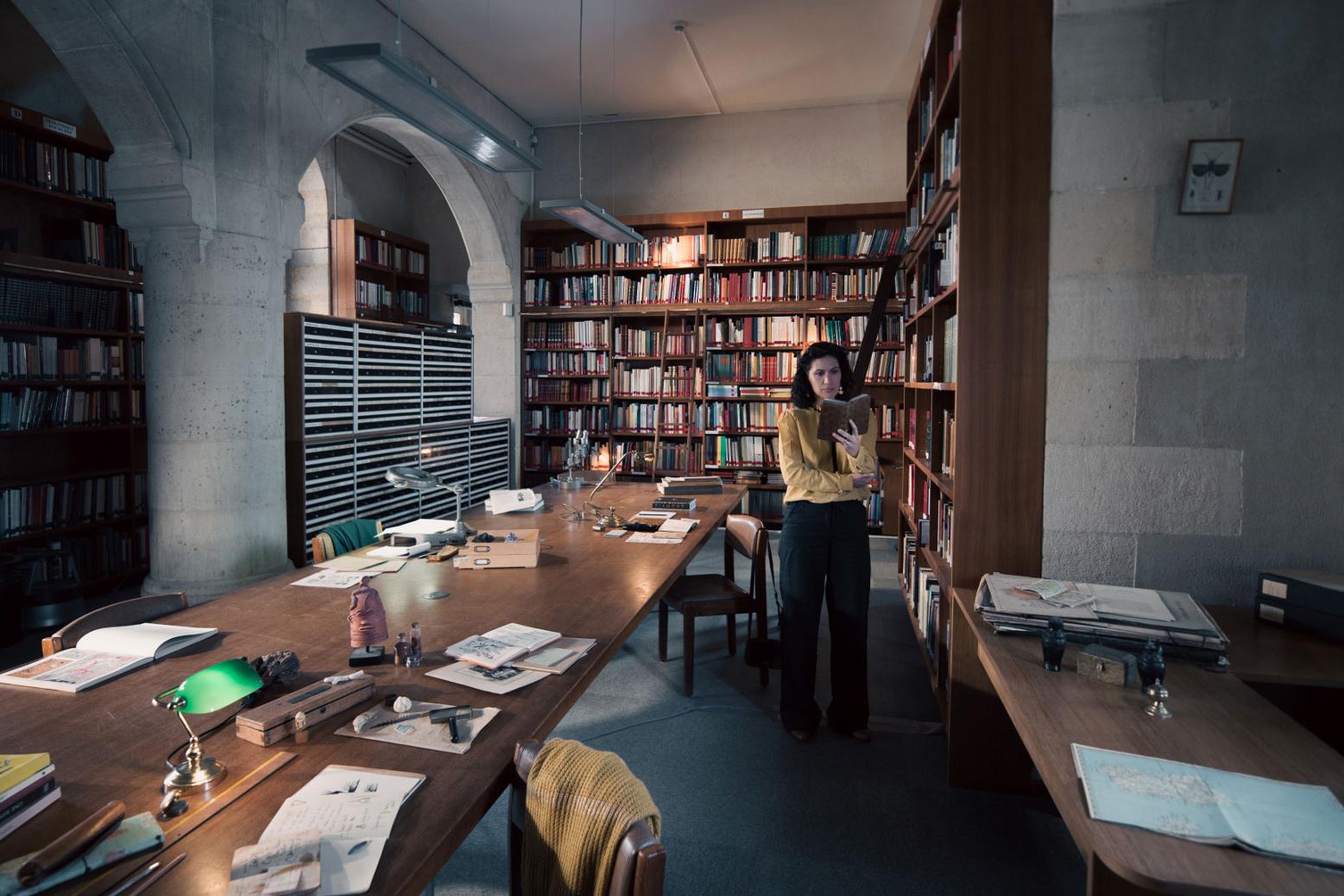 A woman stands in a historic library filled with bookshelves and long wooden tables covered in documents, viewed through a ZEISS lens.