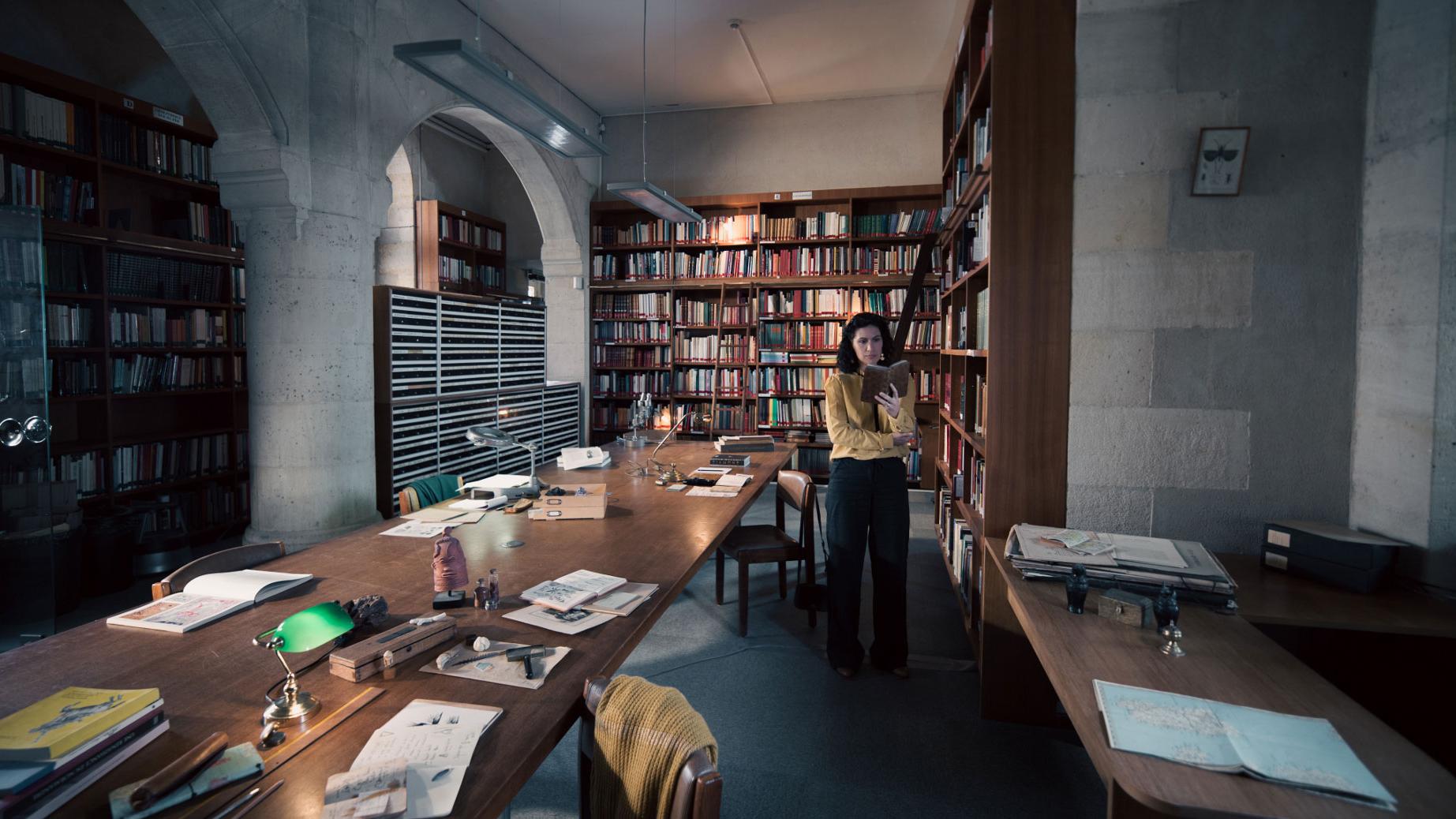 A woman stands in a historic library filled with bookshelves and long wooden tables covered in documents, viewed through a ZEISS lens.