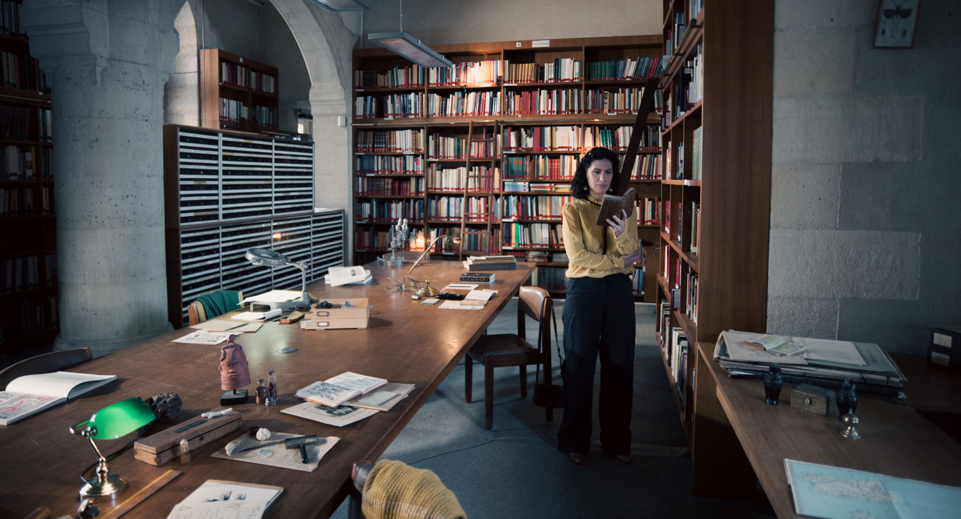 A woman stands in a historic library filled with bookshelves and long wooden tables covered in documents, viewed through a ZEISS lens.