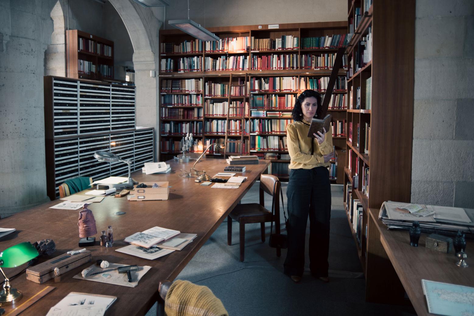 A woman stands in a historic library filled with bookshelves and long wooden tables covered in documents, viewed through a ZEISS lens.