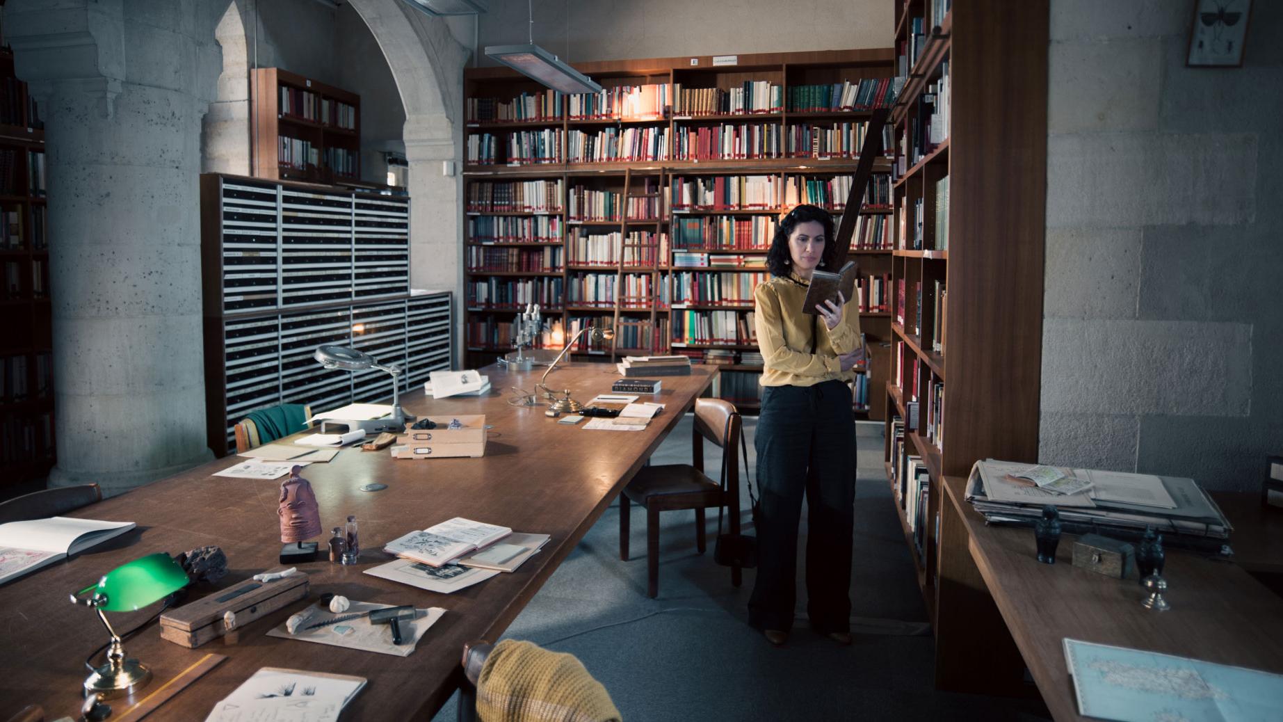 A woman stands in a historic library filled with bookshelves and long wooden tables covered in documents, viewed through a ZEISS lens.