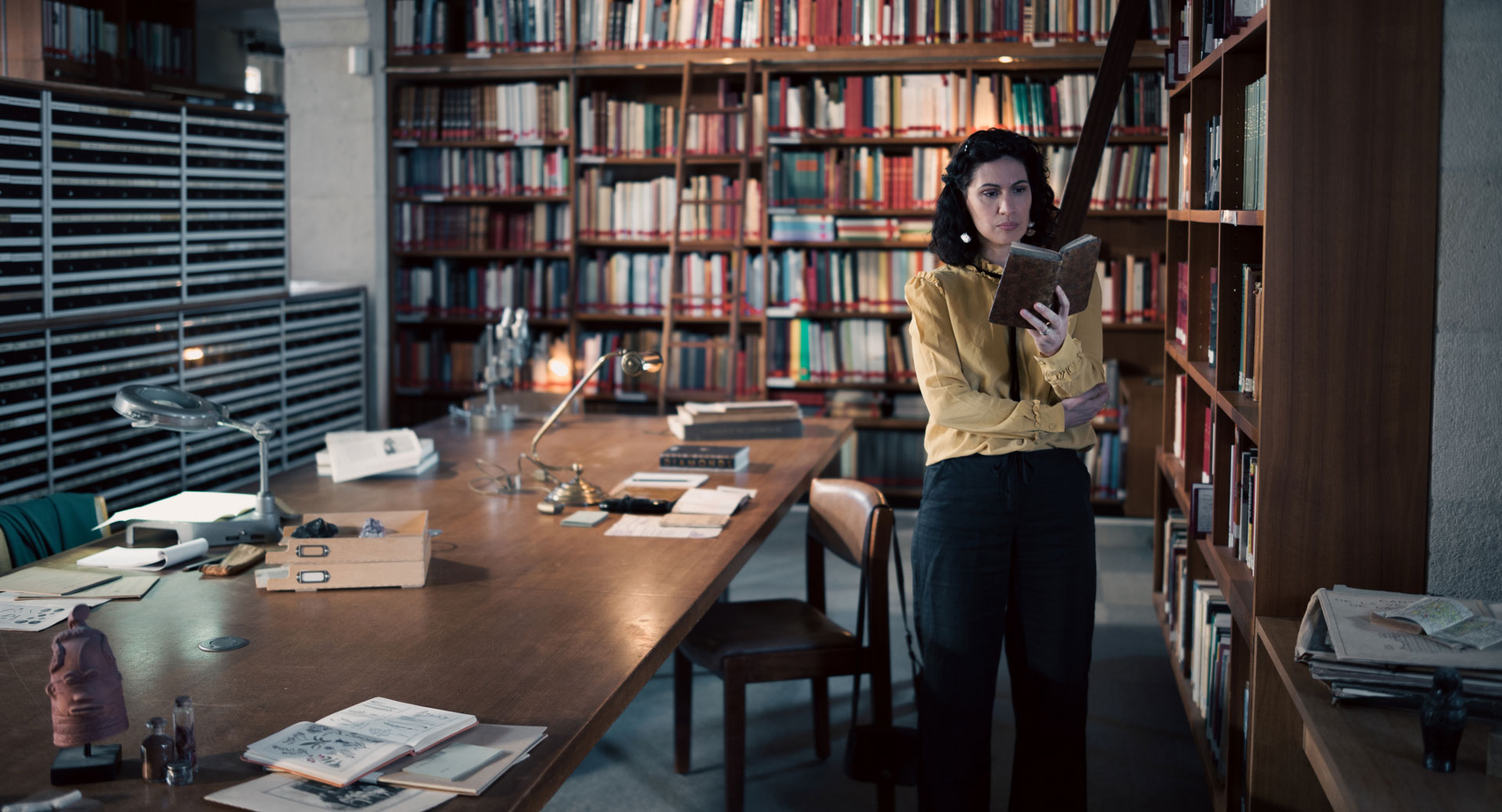 A woman stands in a historic library filled with bookshelves and long wooden tables covered in documents, viewed through a ZEISS lens.