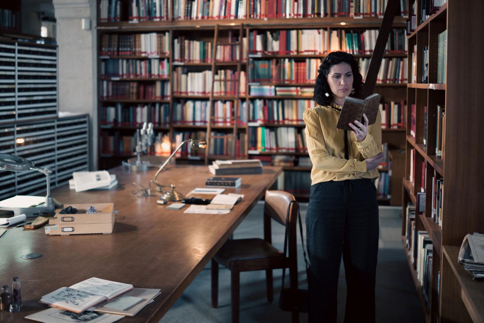 A woman stands in a historic library filled with bookshelves and long wooden tables covered in documents, viewed through a ZEISS lens.