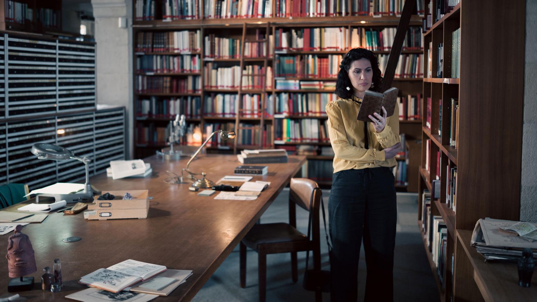 A woman stands in a historic library filled with bookshelves and long wooden tables covered in documents, viewed through a ZEISS lens.