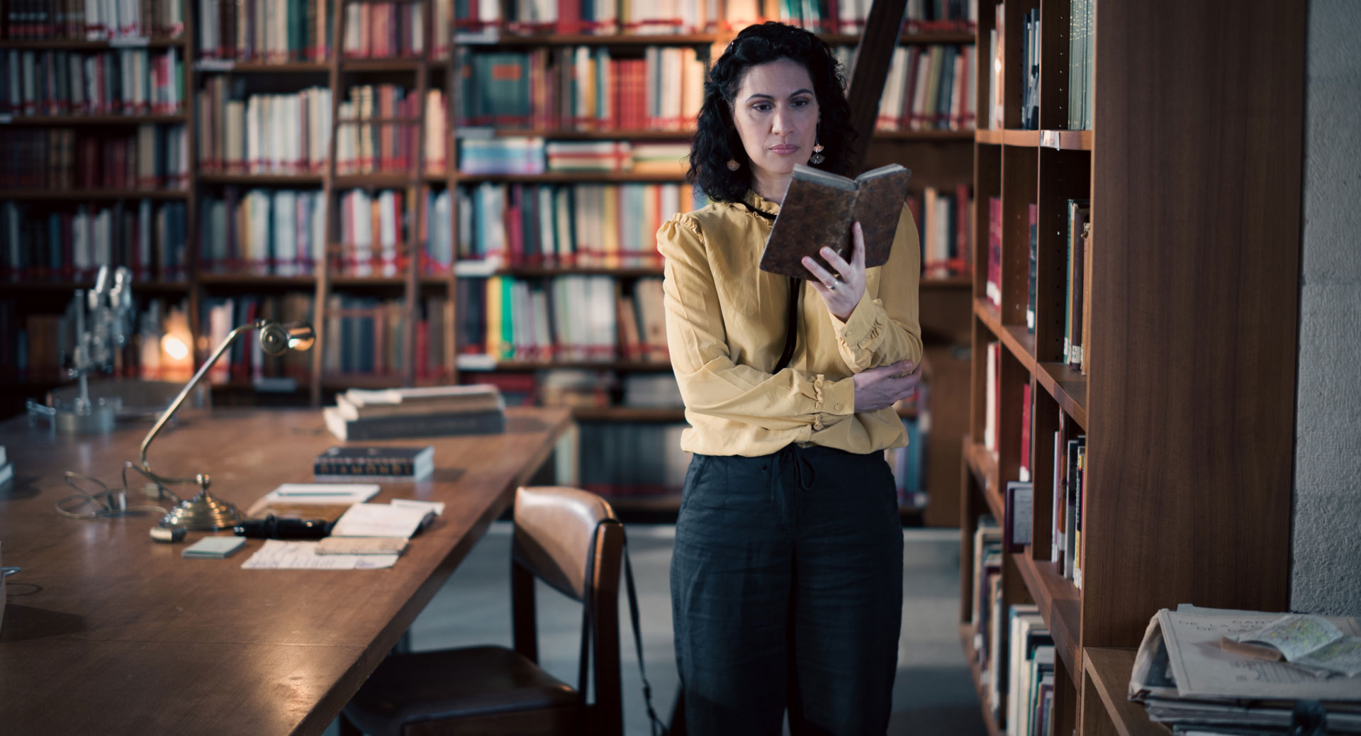 A woman stands in a historic library filled with bookshelves and long wooden tables covered in documents, viewed through a ZEISS lens.