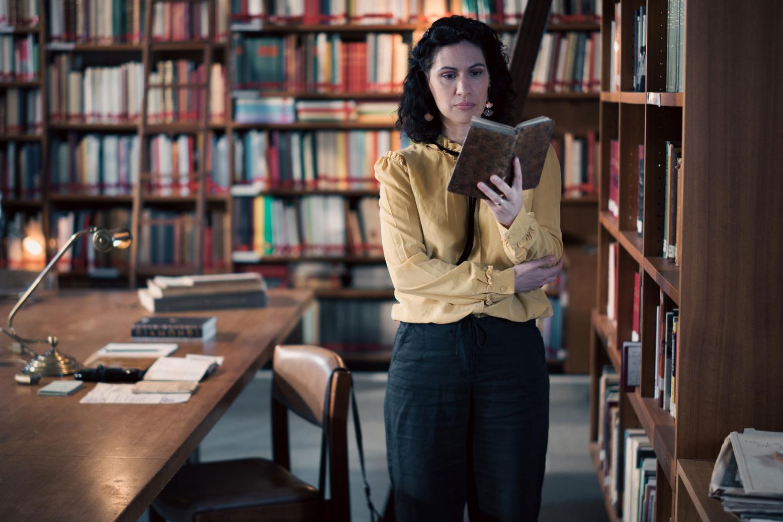 A woman stands in a historic library filled with bookshelves and long wooden tables covered in documents, viewed through a ZEISS lens.