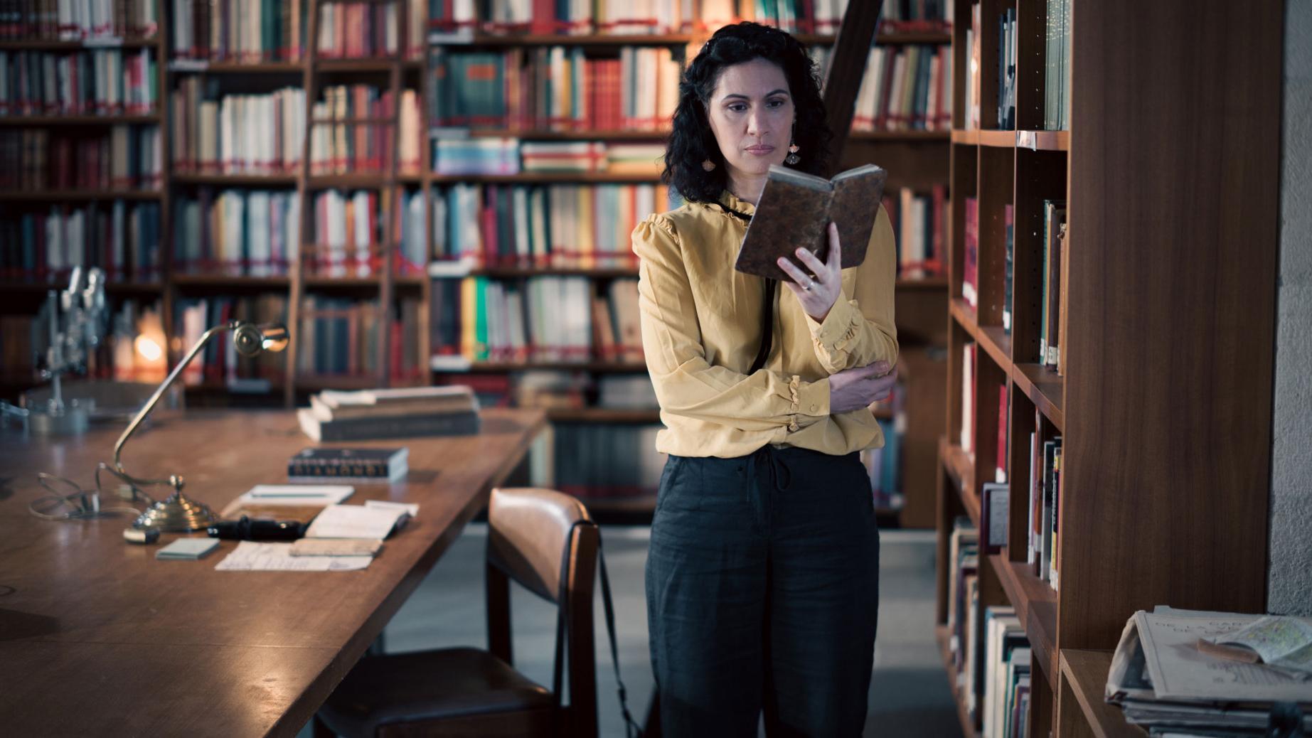A woman stands in a historic library filled with bookshelves and long wooden tables covered in documents, viewed through a ZEISS lens.