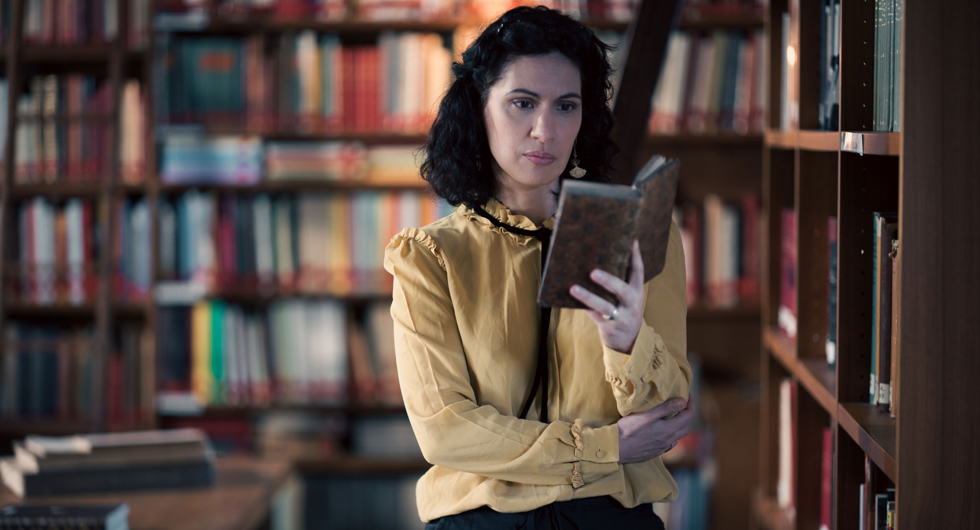 A woman stands in a historic library filled with bookshelves and long wooden tables covered in documents, viewed through a ZEISS lens.