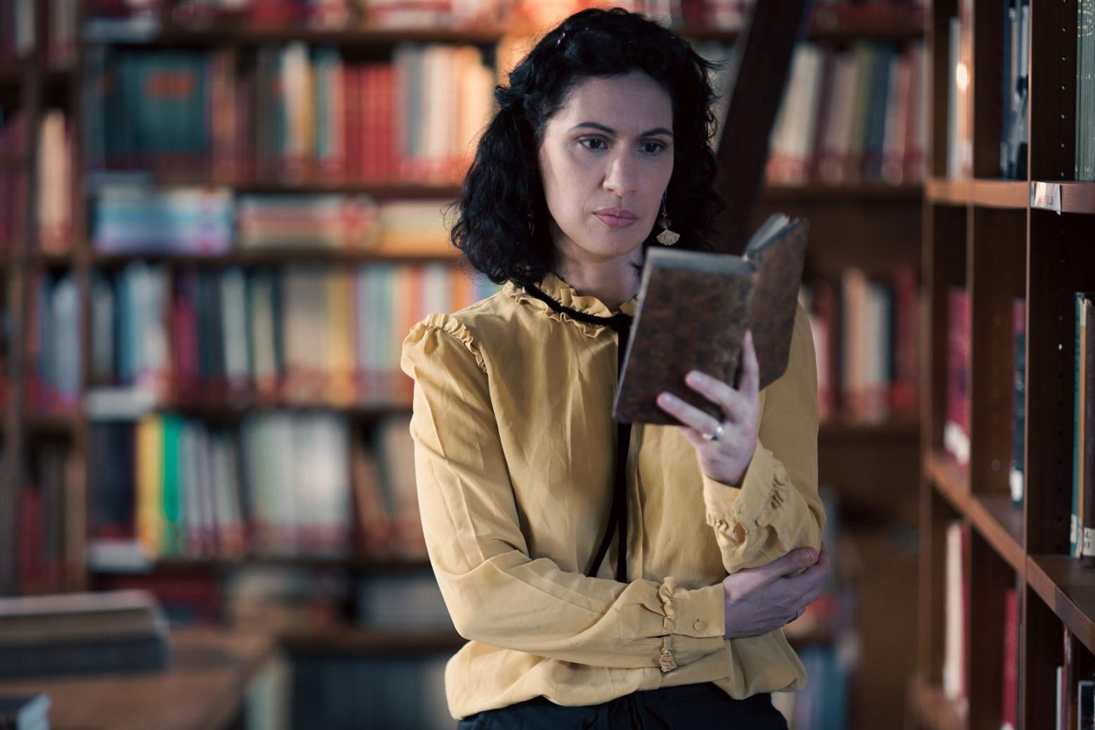 A woman stands in a historic library filled with bookshelves and long wooden tables covered in documents, viewed through a ZEISS lens.