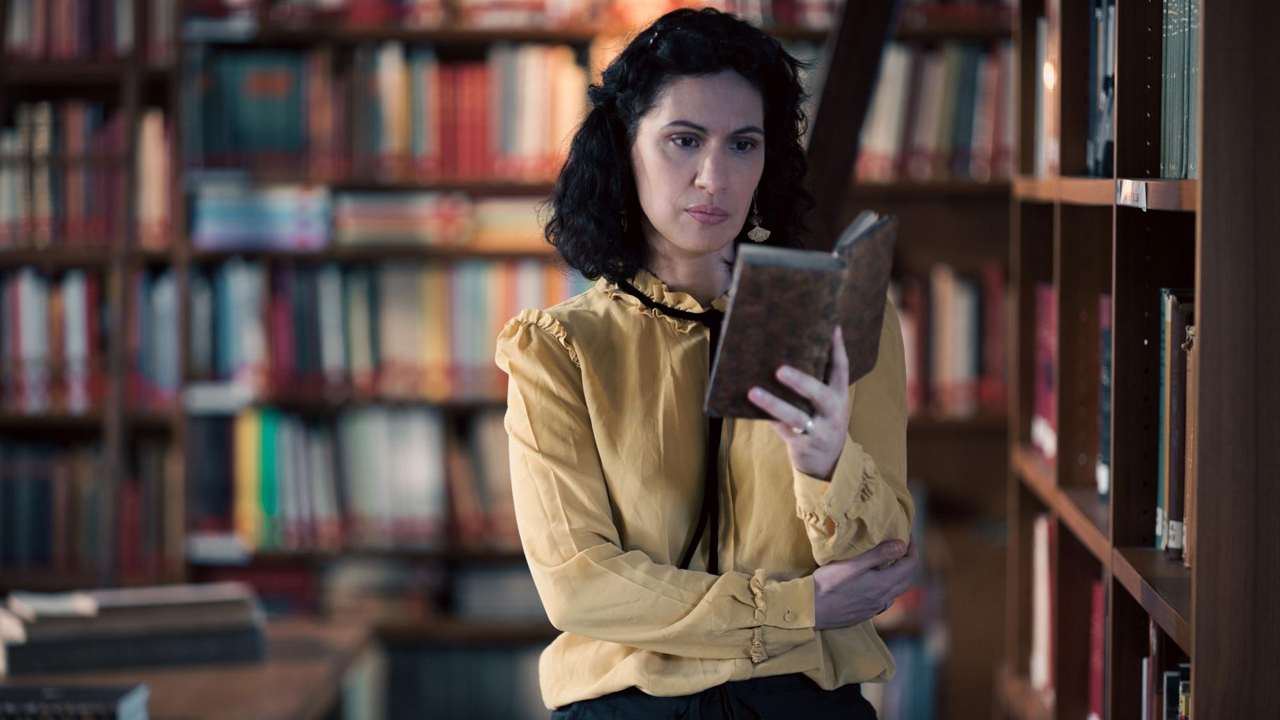 A woman stands in a historic library filled with bookshelves and long wooden tables covered in documents, viewed through a ZEISS lens.
