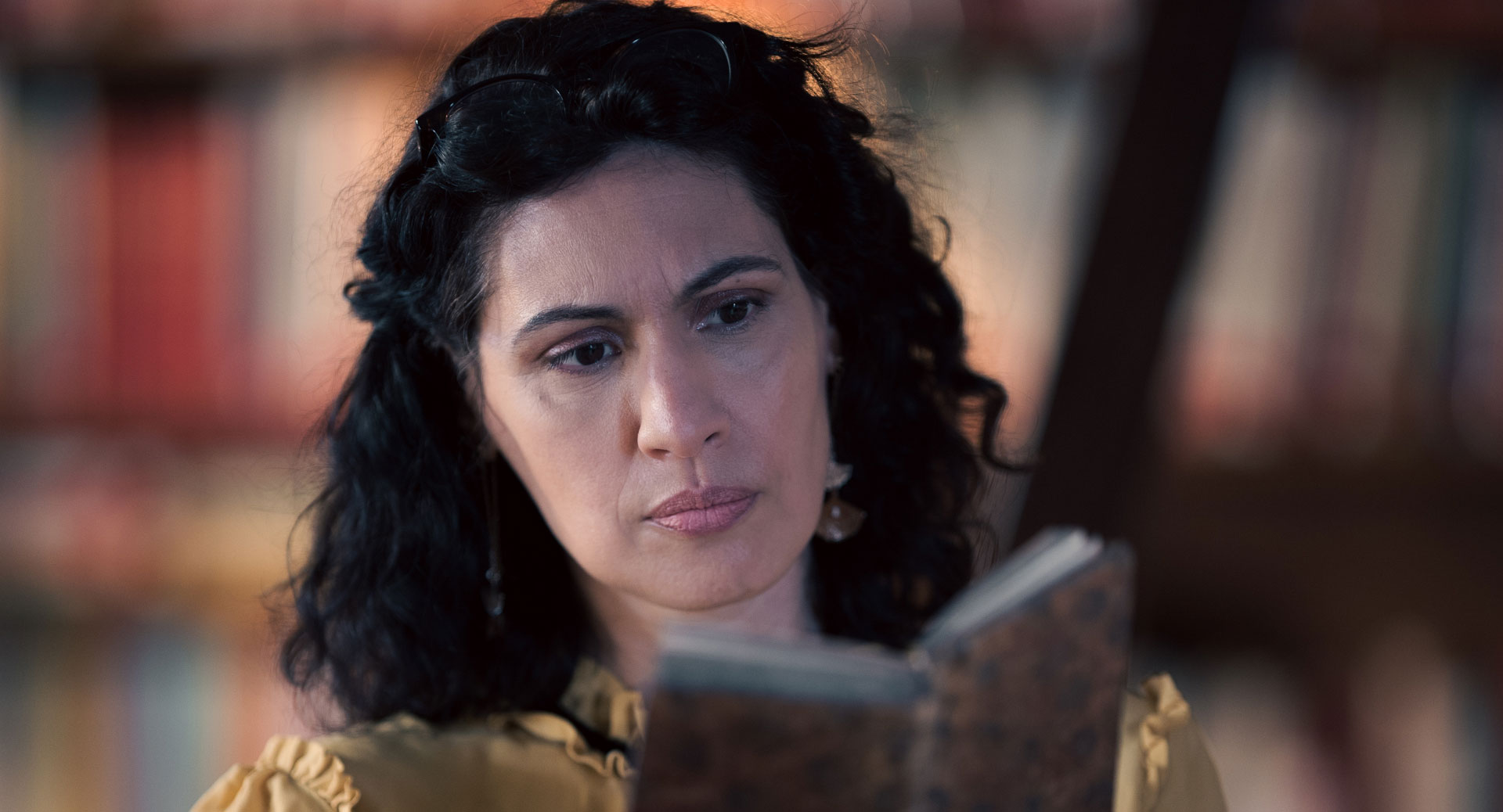 A woman stands in a historic library filled with bookshelves and long wooden tables covered in documents, viewed through a ZEISS lens.