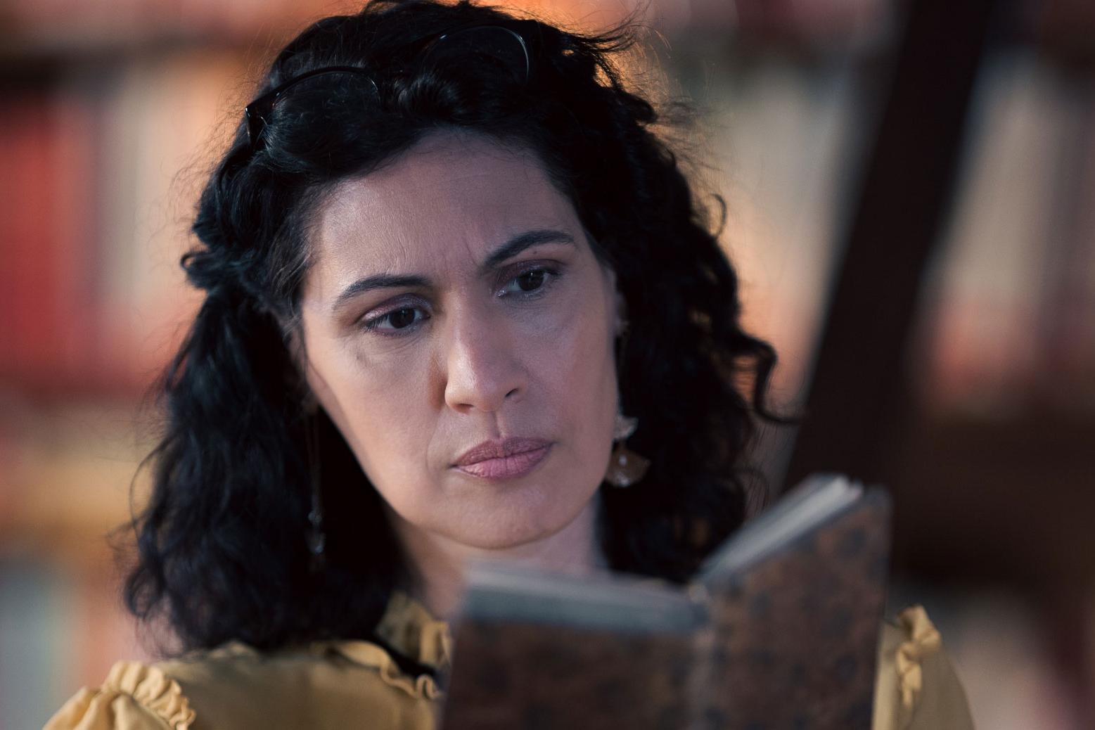 A woman stands in a historic library filled with bookshelves and long wooden tables covered in documents, viewed through a ZEISS lens.