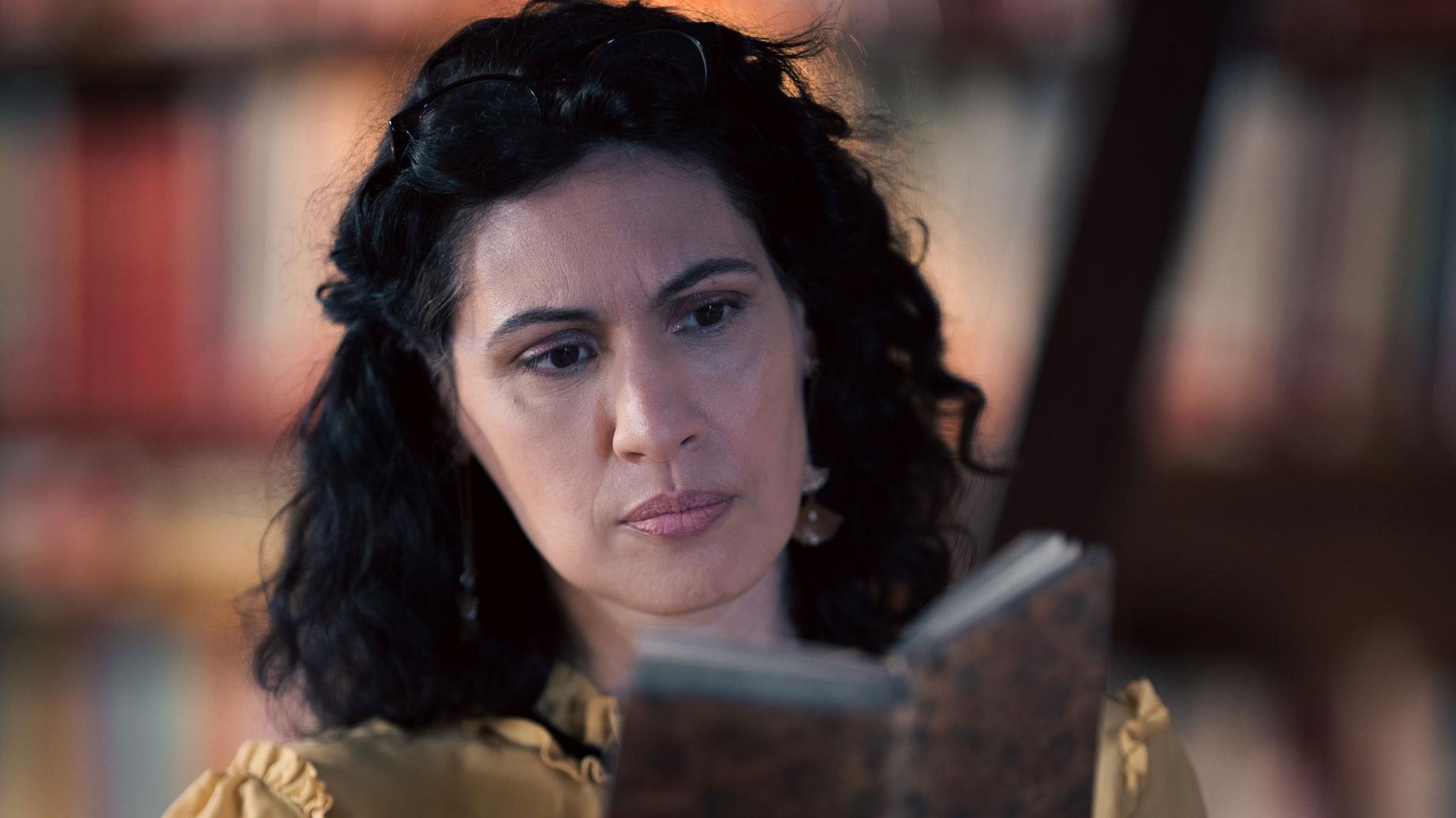 A woman stands in a historic library filled with bookshelves and long wooden tables covered in documents, viewed through a ZEISS lens.