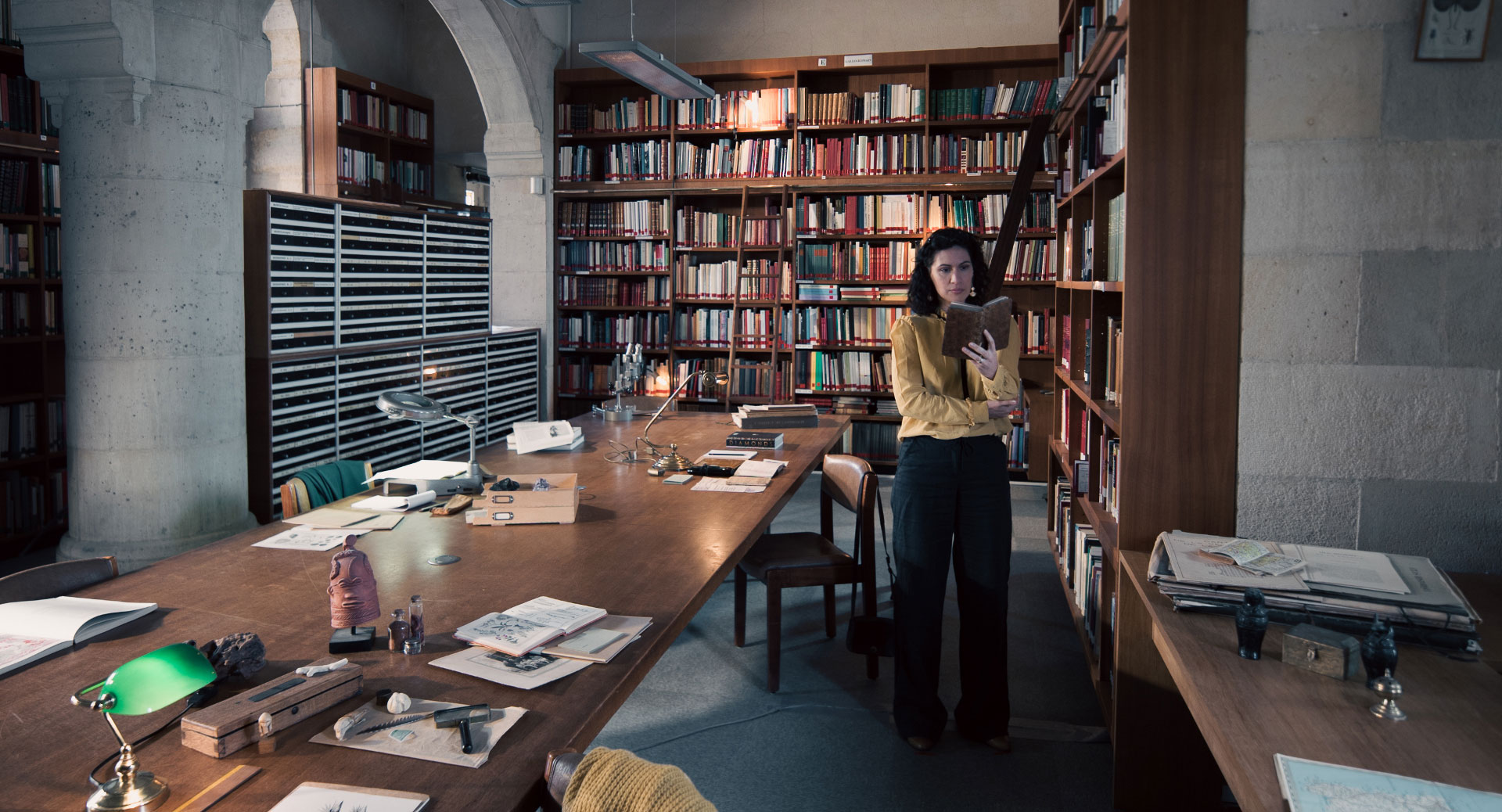 A woman stands in a historic library filled with bookshelves and long wooden tables covered in documents, viewed through a ZEISS lens.