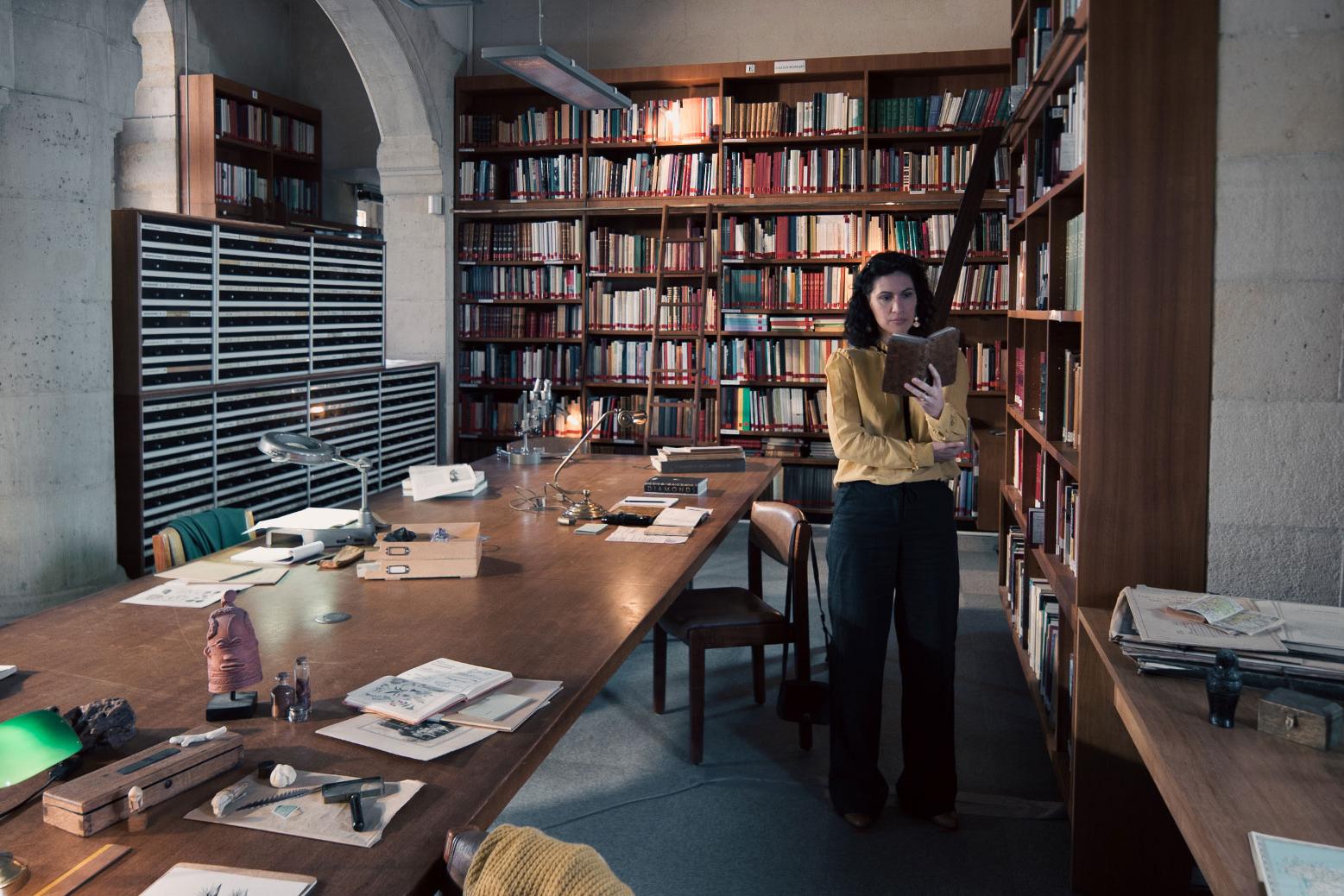 A woman stands in a historic library filled with bookshelves and long wooden tables covered in documents, viewed through a ZEISS lens.