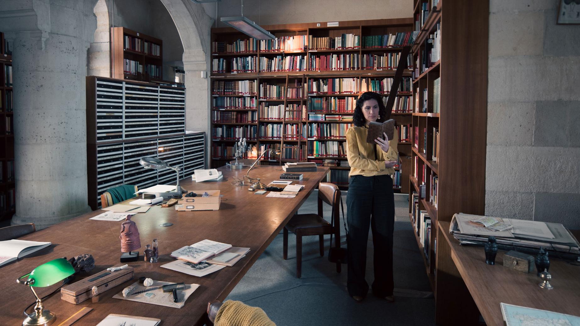 A woman stands in a historic library filled with bookshelves and long wooden tables covered in documents, viewed through a ZEISS lens.