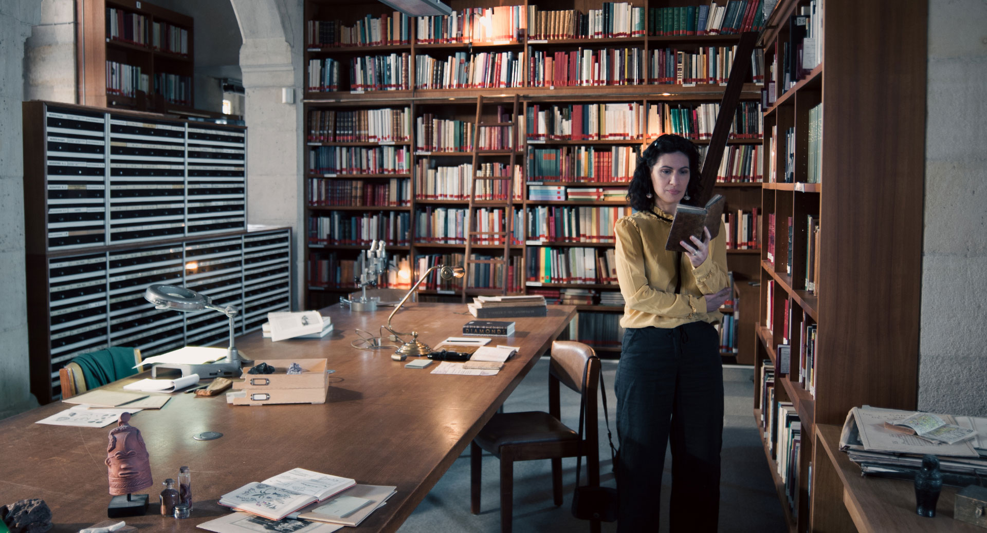 A woman stands in a historic library filled with bookshelves and long wooden tables covered in documents, viewed through a ZEISS lens.