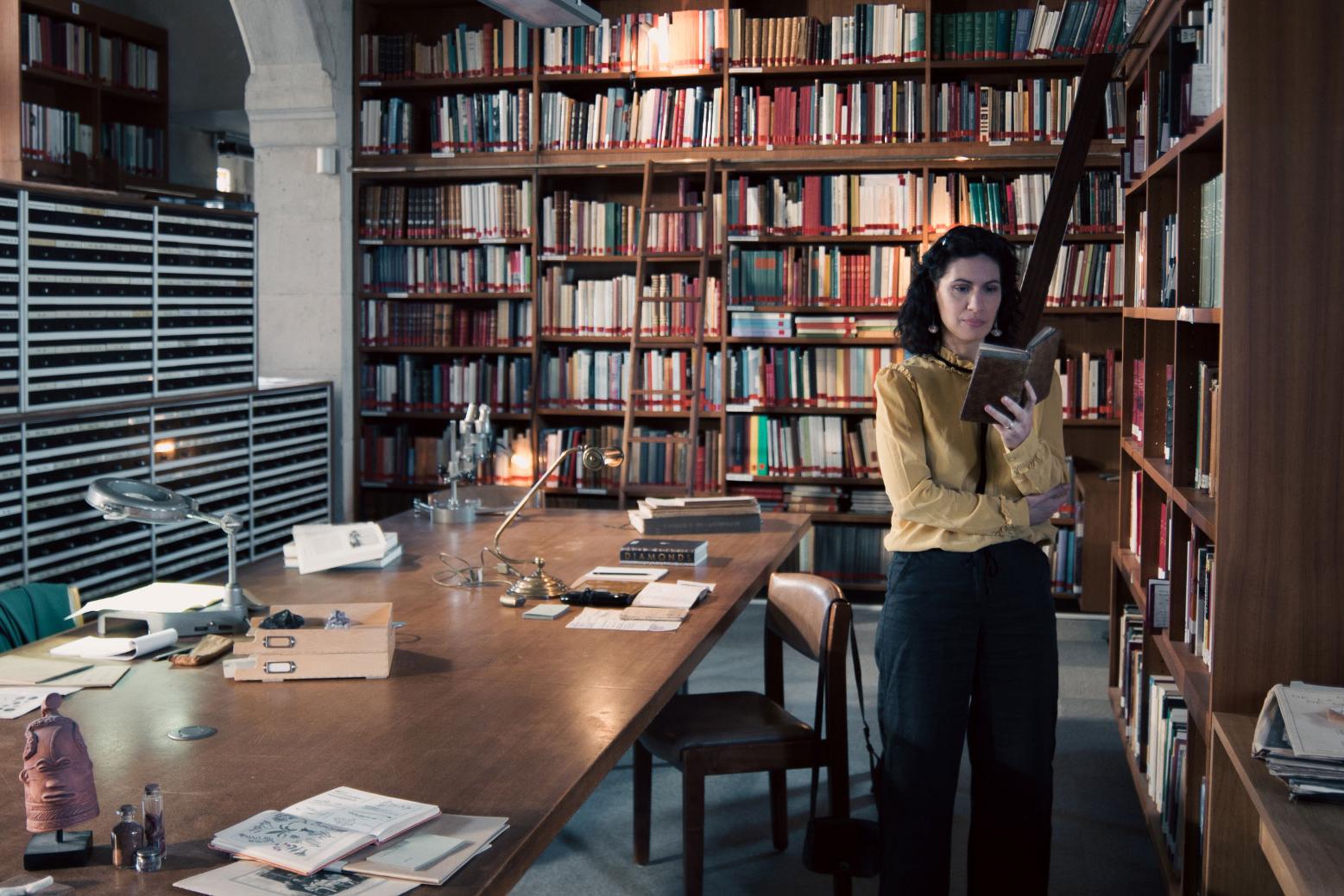 A woman stands in a historic library filled with bookshelves and long wooden tables covered in documents, viewed through a ZEISS lens.