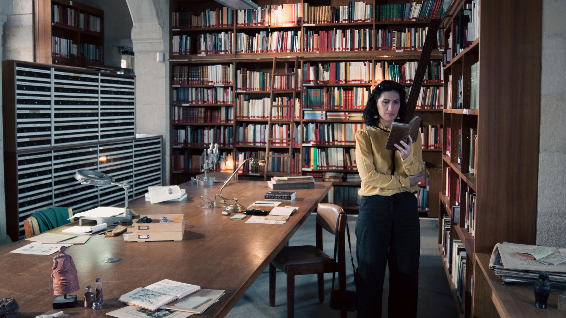 A woman stands in a historic library filled with bookshelves and long wooden tables covered in documents, viewed through a ZEISS lens.