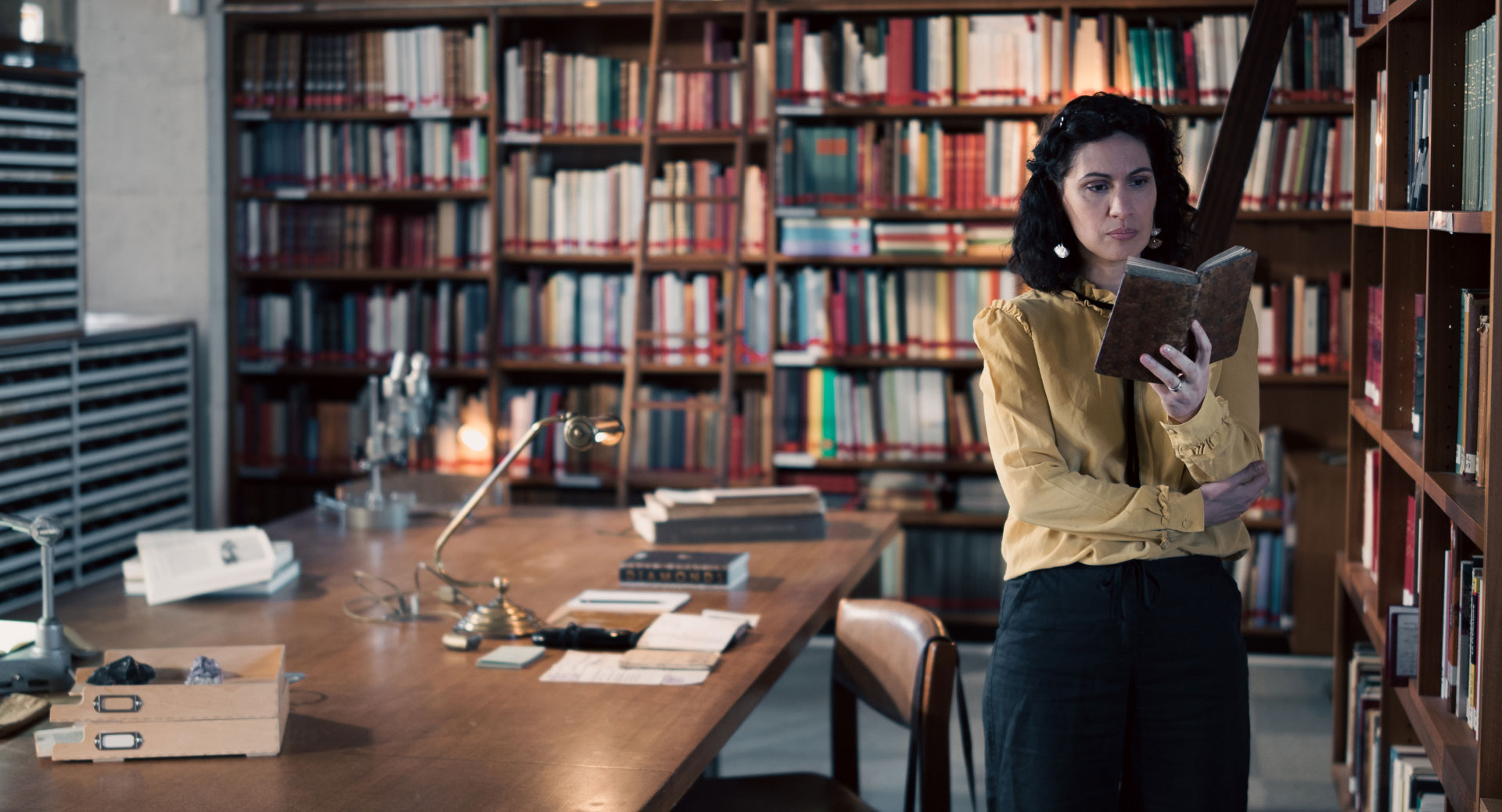 A woman stands in a historic library filled with bookshelves and long wooden tables covered in documents, viewed through a ZEISS lens.