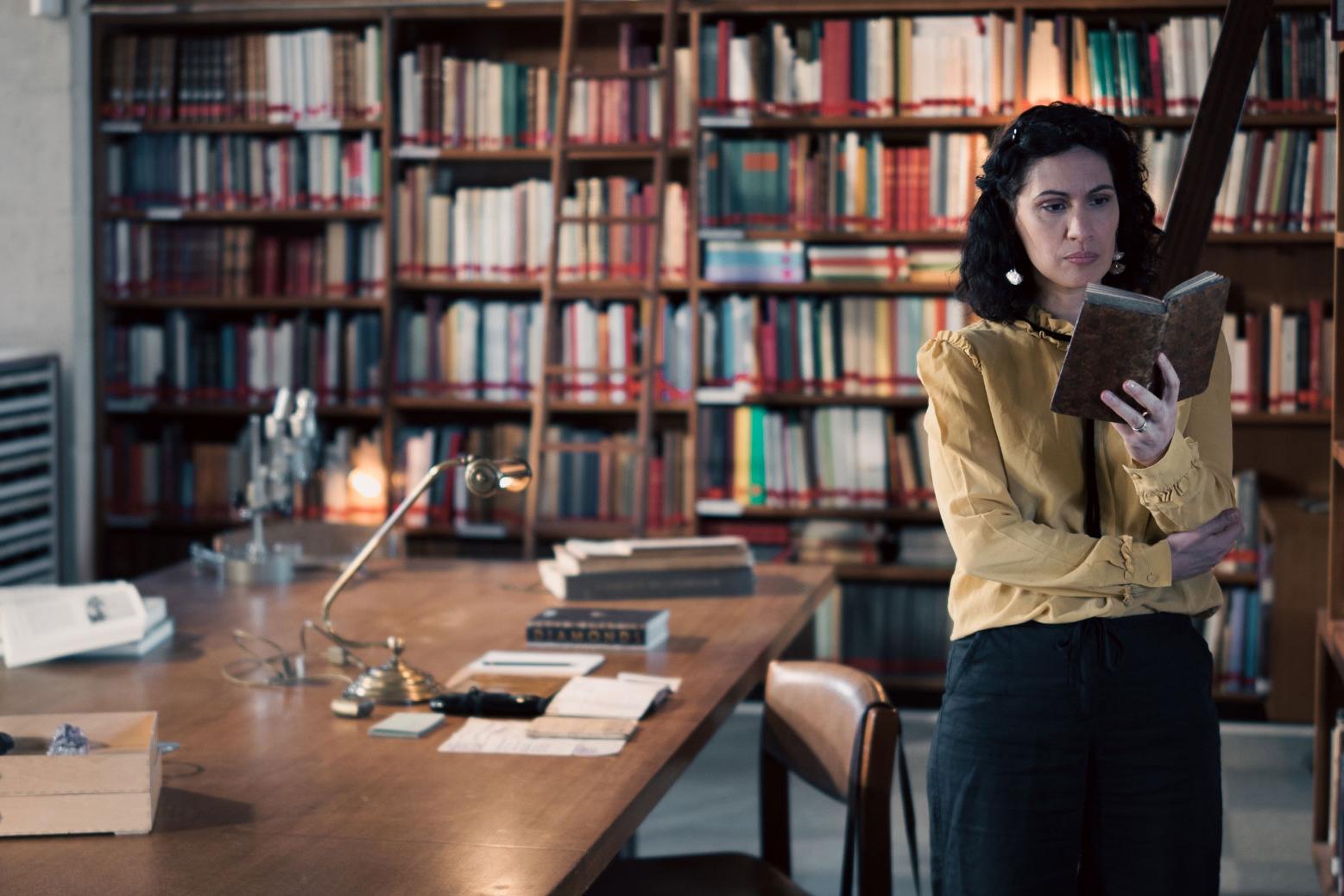 A woman stands in a historic library filled with bookshelves and long wooden tables covered in documents, viewed through a ZEISS lens.
