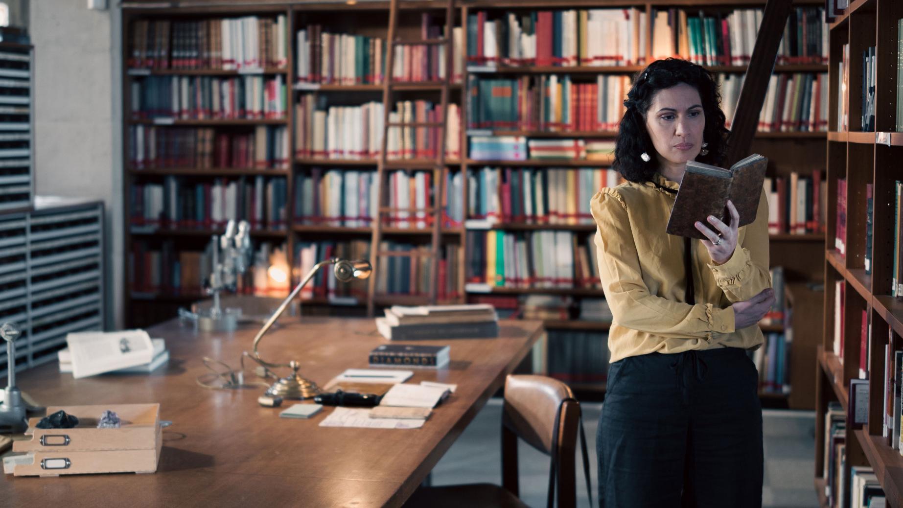 A woman stands in a historic library filled with bookshelves and long wooden tables covered in documents, viewed through a ZEISS lens.