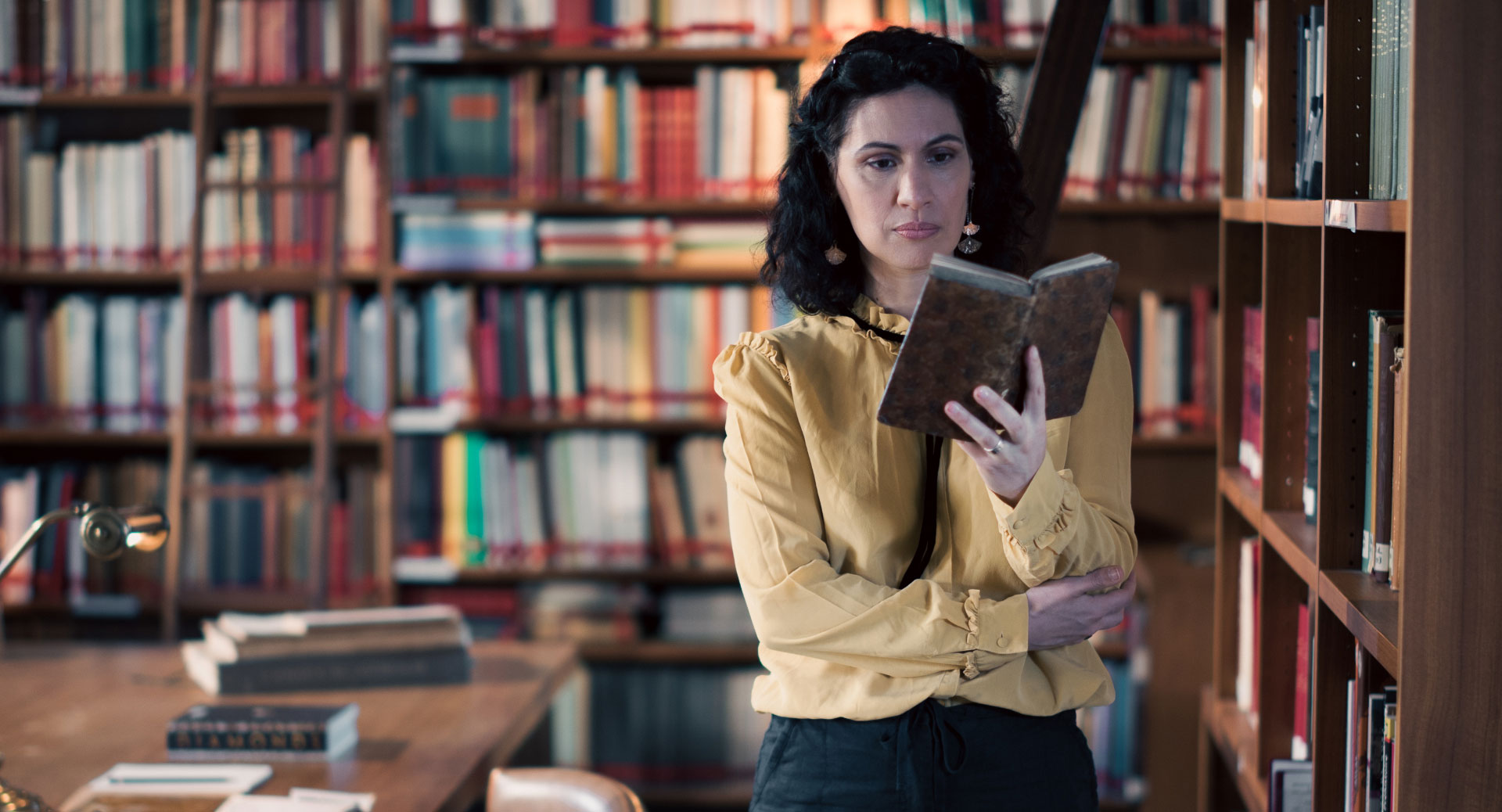 A woman stands in a historic library filled with bookshelves and long wooden tables covered in documents, viewed through a ZEISS lens.