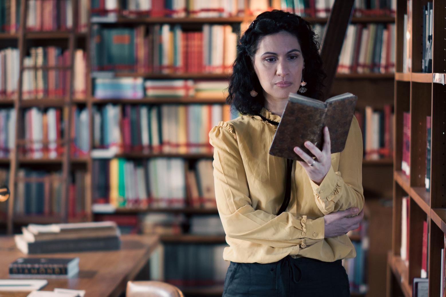 A woman stands in a historic library filled with bookshelves and long wooden tables covered in documents, viewed through a ZEISS lens.