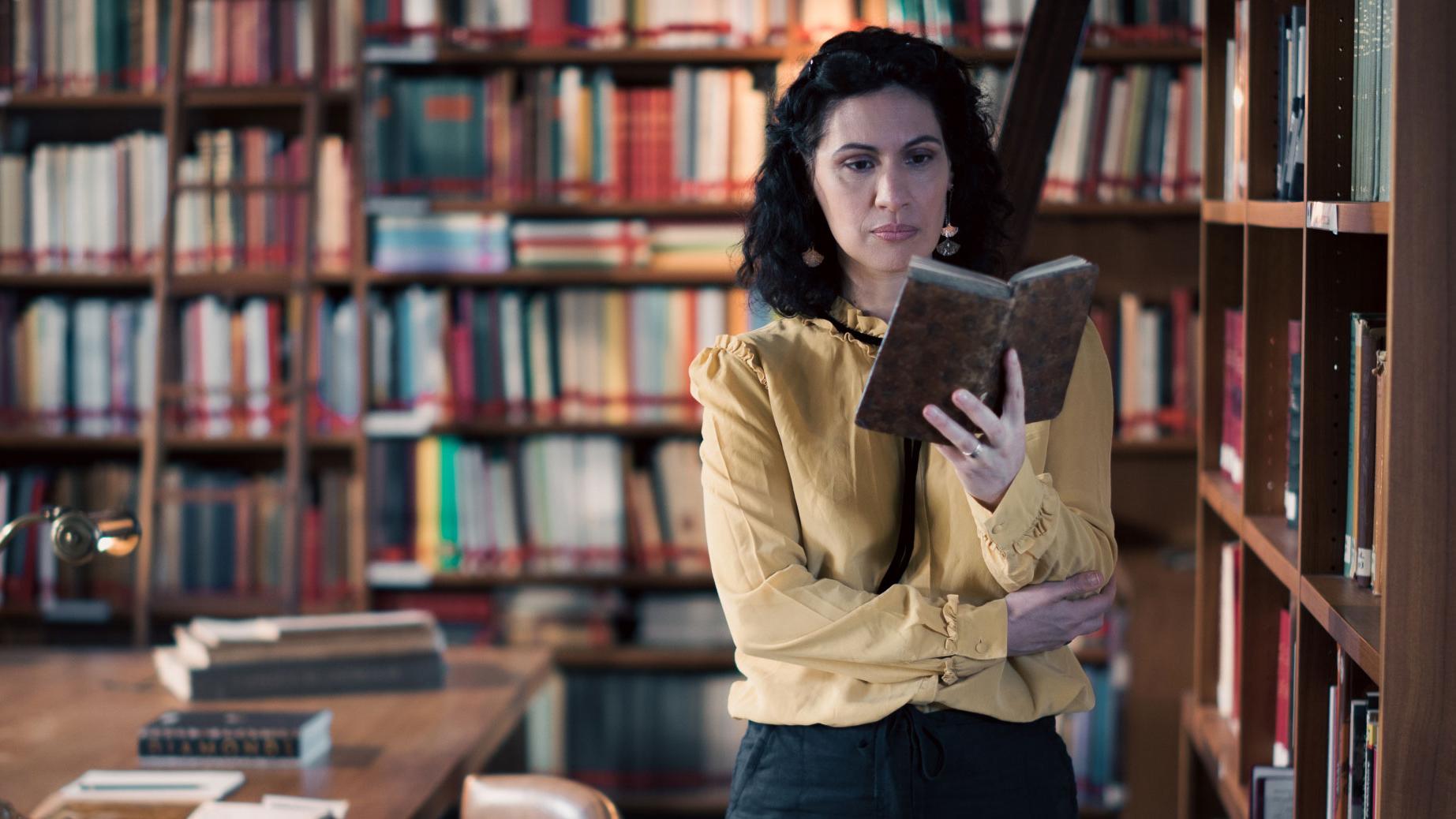 A woman stands in a historic library filled with bookshelves and long wooden tables covered in documents, viewed through a ZEISS lens.