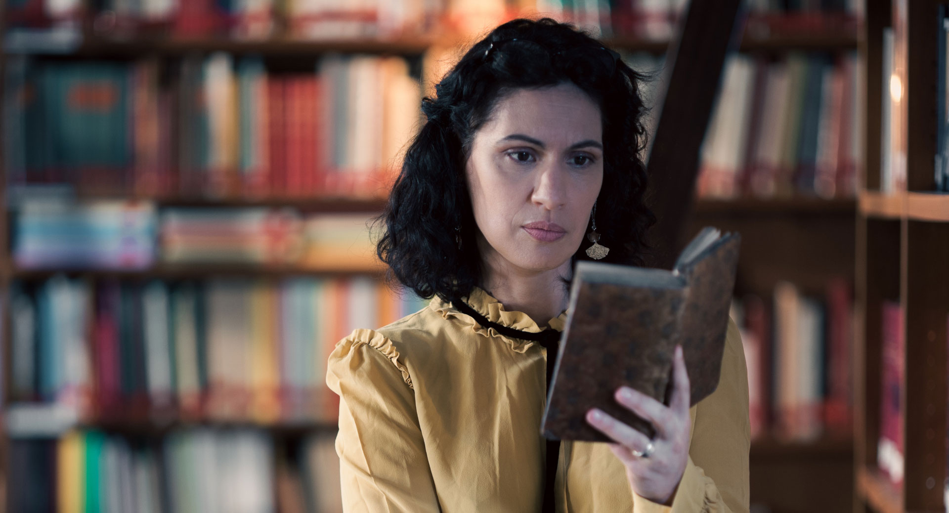 A woman stands in a historic library filled with bookshelves and long wooden tables covered in documents, viewed through a ZEISS lens.