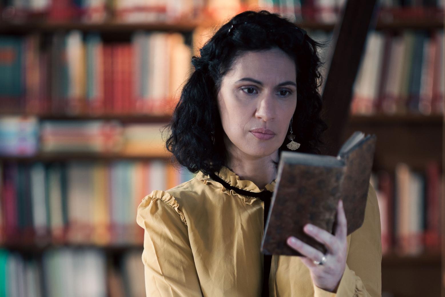 A woman stands in a historic library filled with bookshelves and long wooden tables covered in documents, viewed through a ZEISS lens.